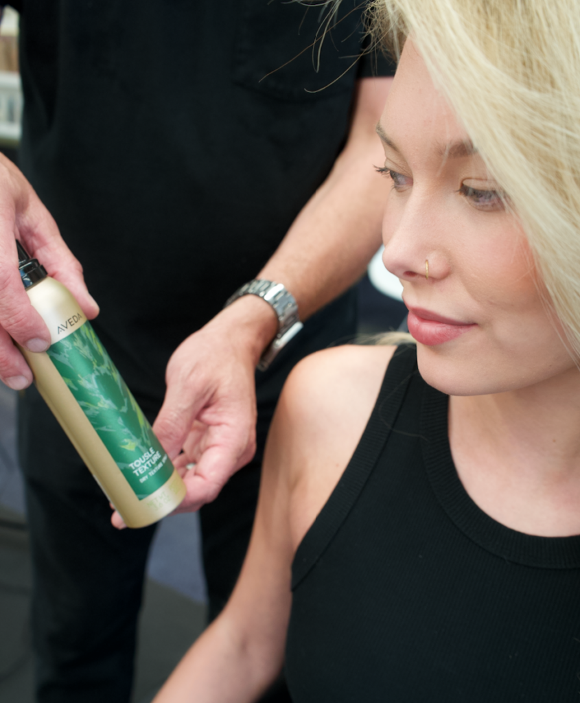 A bottle of Aveda Tousle Texture Dry Spray being used to create effortless, piecey volume on a model with soft waves.
