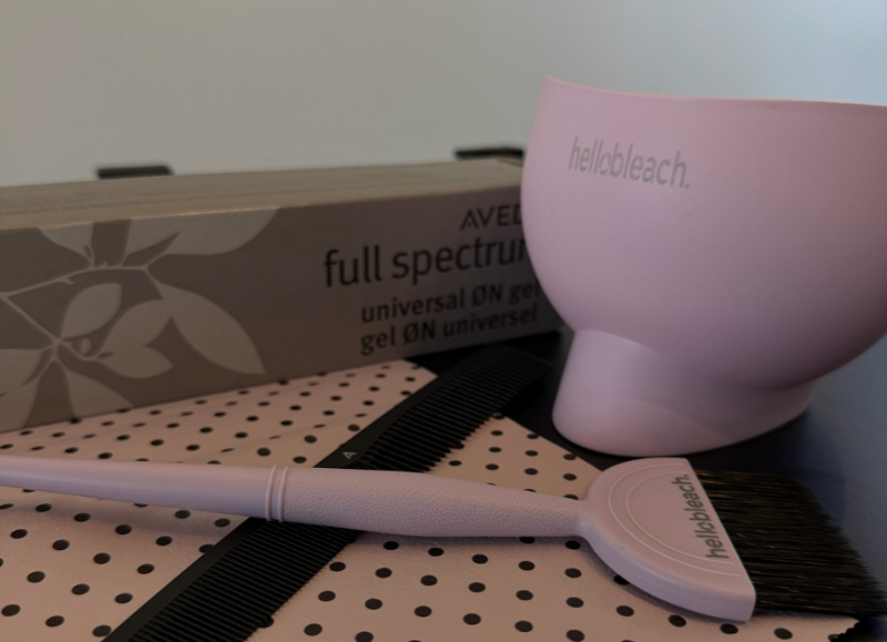 A pastel pink Hello Bleach sustainable salon tools set, featuring an ergonomic tint bowl and color brush resting next to a box of Aveda Full Spectrum hair color and a black cutting comb.