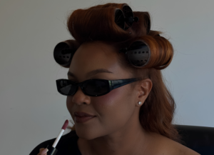 A model with vibrant auburn hair set in large black volume rollers, wearing dark sunglasses and applying lip gloss during a behind-the-scenes salon content capture photoshoot.