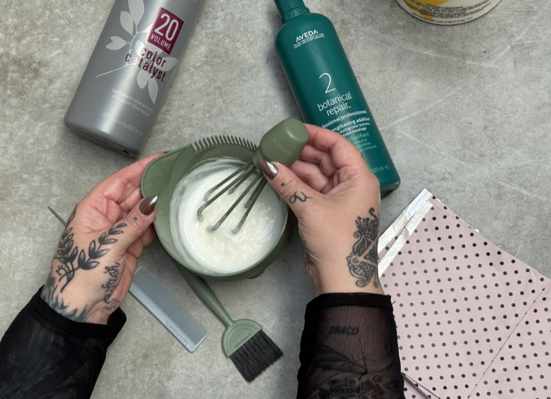 Overhead view of a tattooed stylist using a sage green Hello Bleach whisk to mix hair color in a matching green tint bowl, flanked by Aveda Botanical Repair and Color Catalyst bottles.