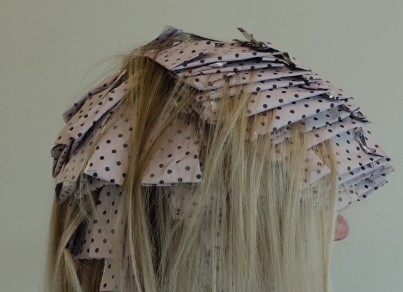 Close-up rear view of a blonde model’s hair neatly sectioned and packaged in Hello Bleach signature pink and black polka-dot patterned hair foils during a highlighting service.